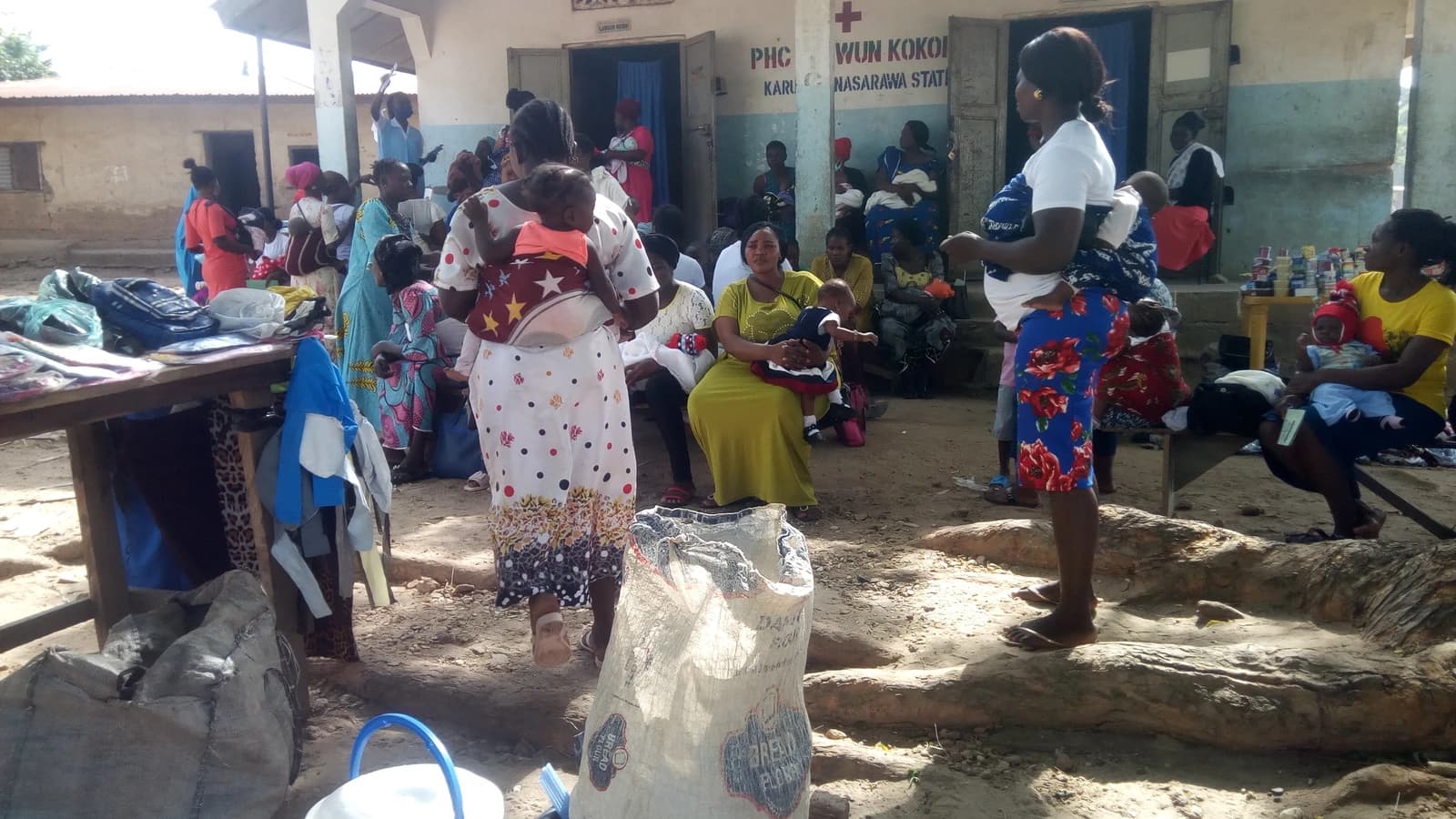 Mothers holding infants wait outside a primary healthcare center in a rural area.