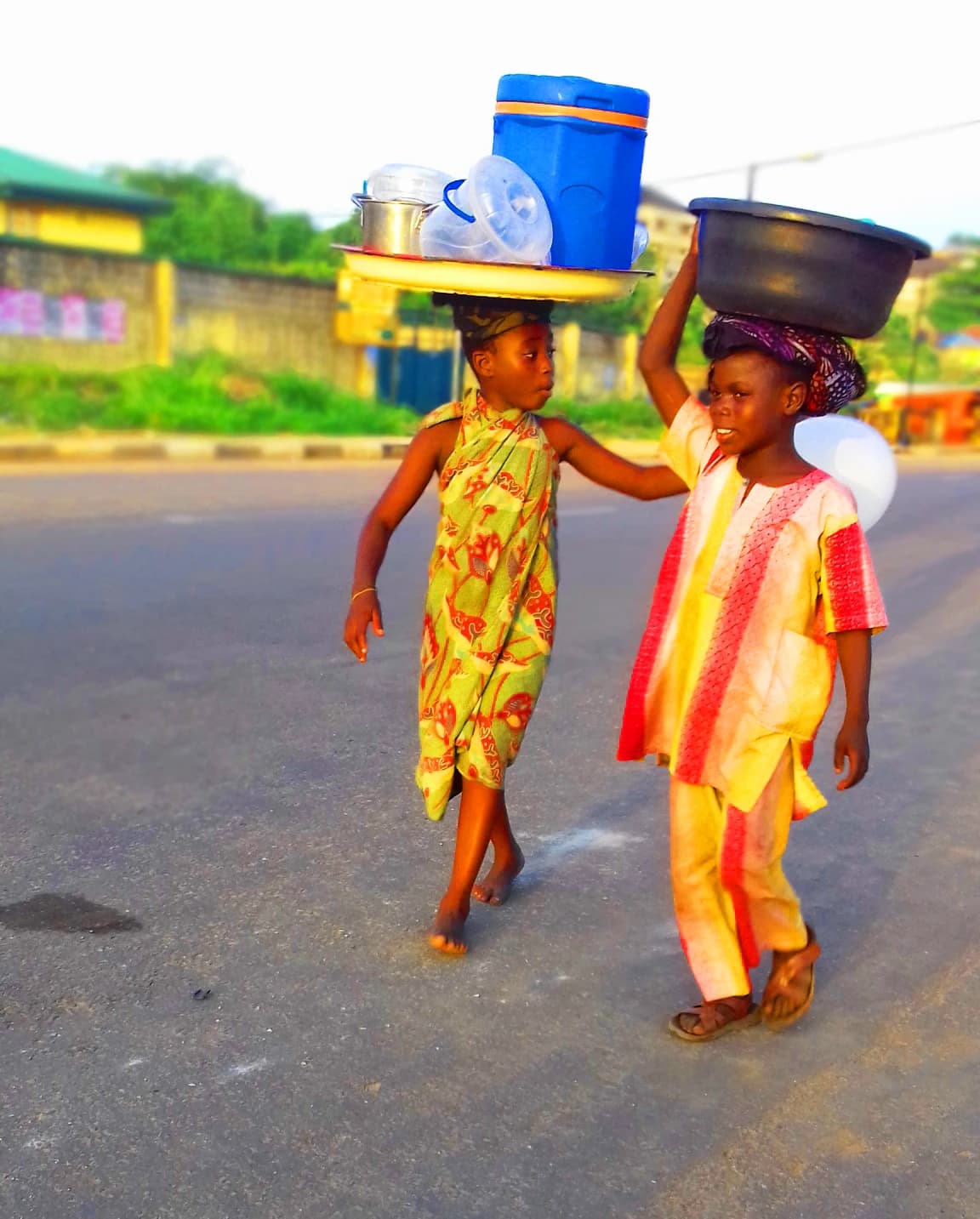 Two Nigerian children carrying items on their heads while walking on a street.