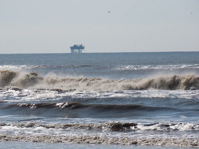 Ocean waves in the foreground with an offshore oil rig visible in the distance.
