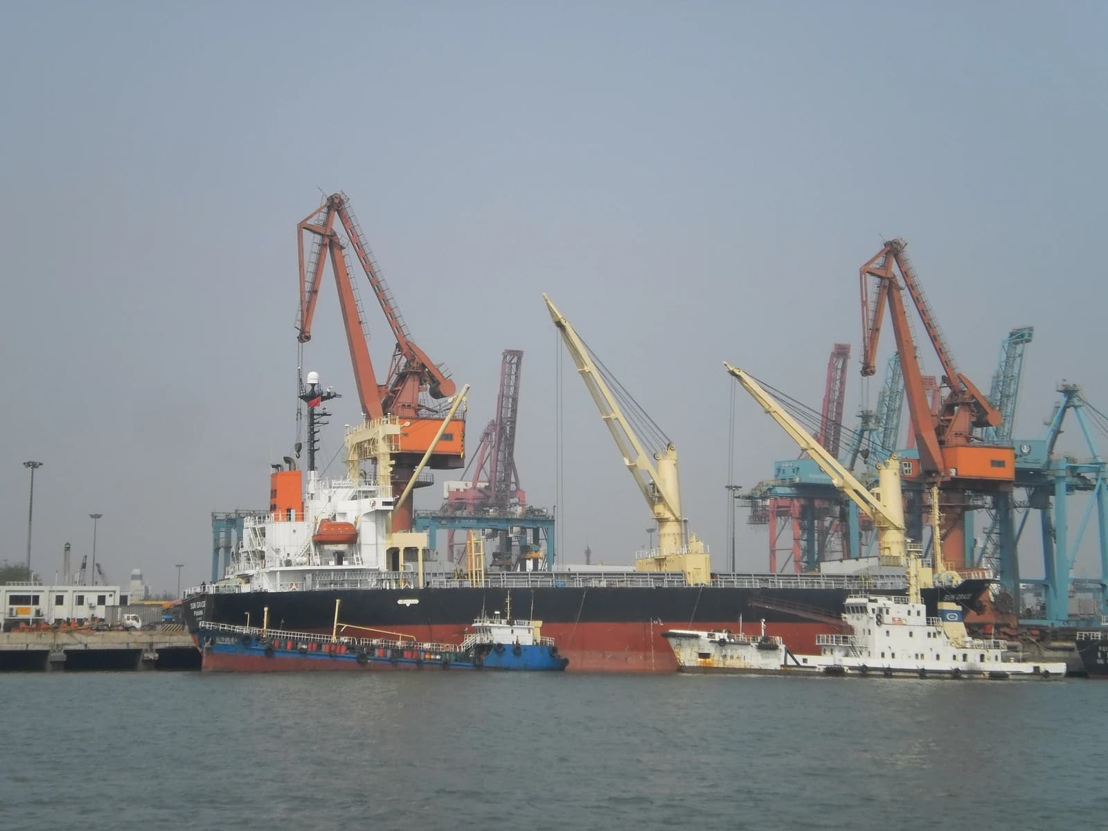 Bunker barges supplying a ship at a port with large cranes in the background.