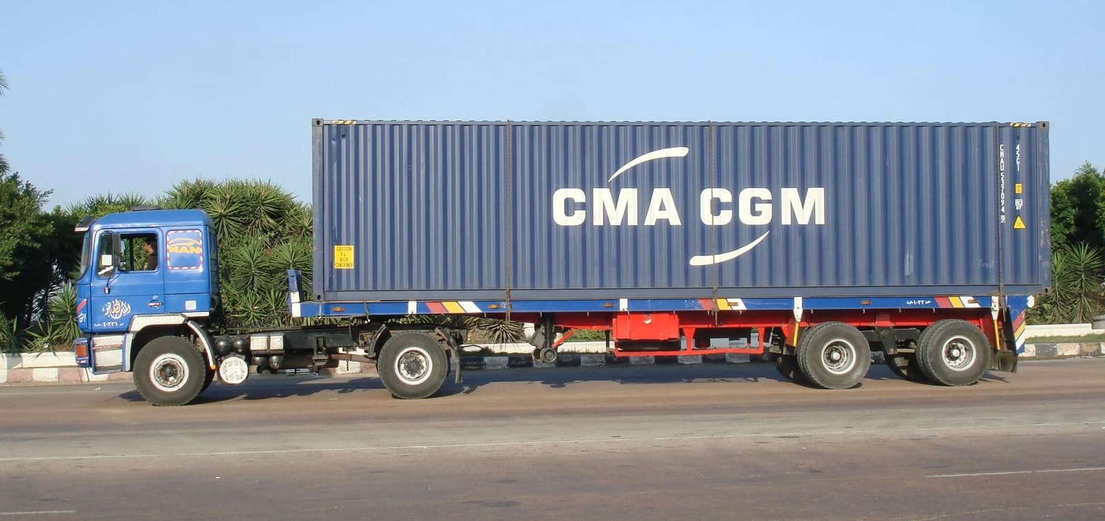 A blue CMA CGM shipping container being transported on a flatbed truck on a road.