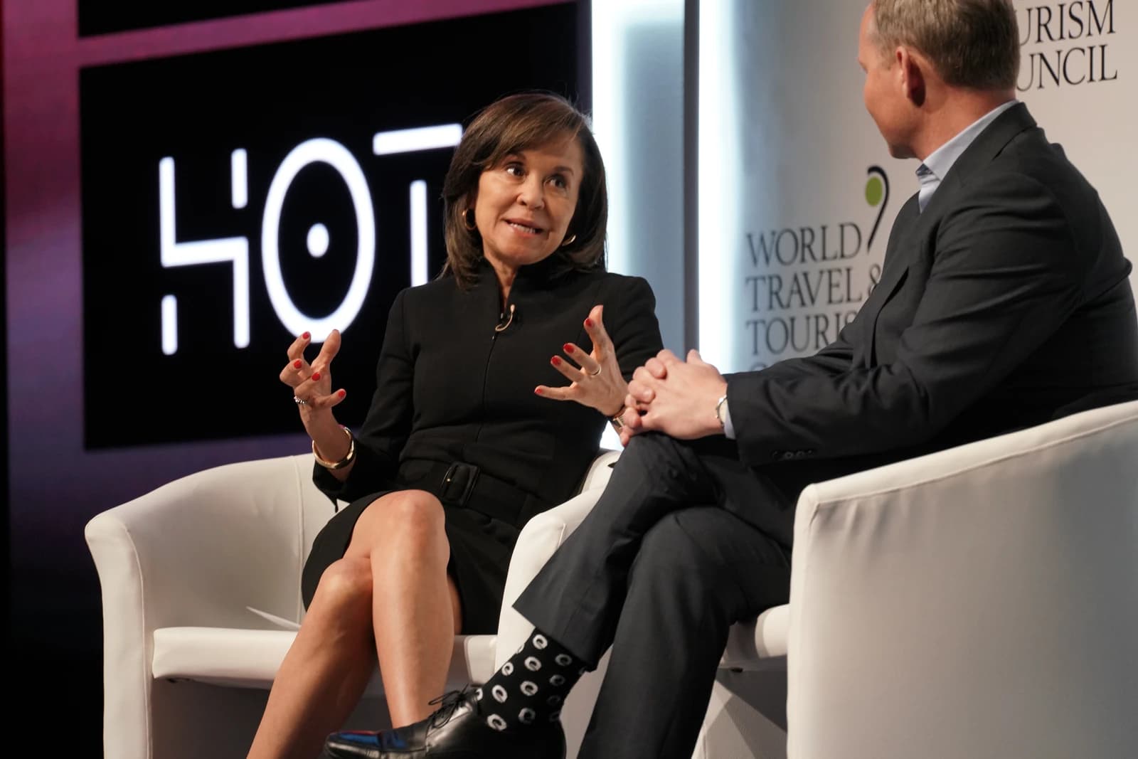 Woman with brown hair speaking, gesturing with hands, seated next to man.