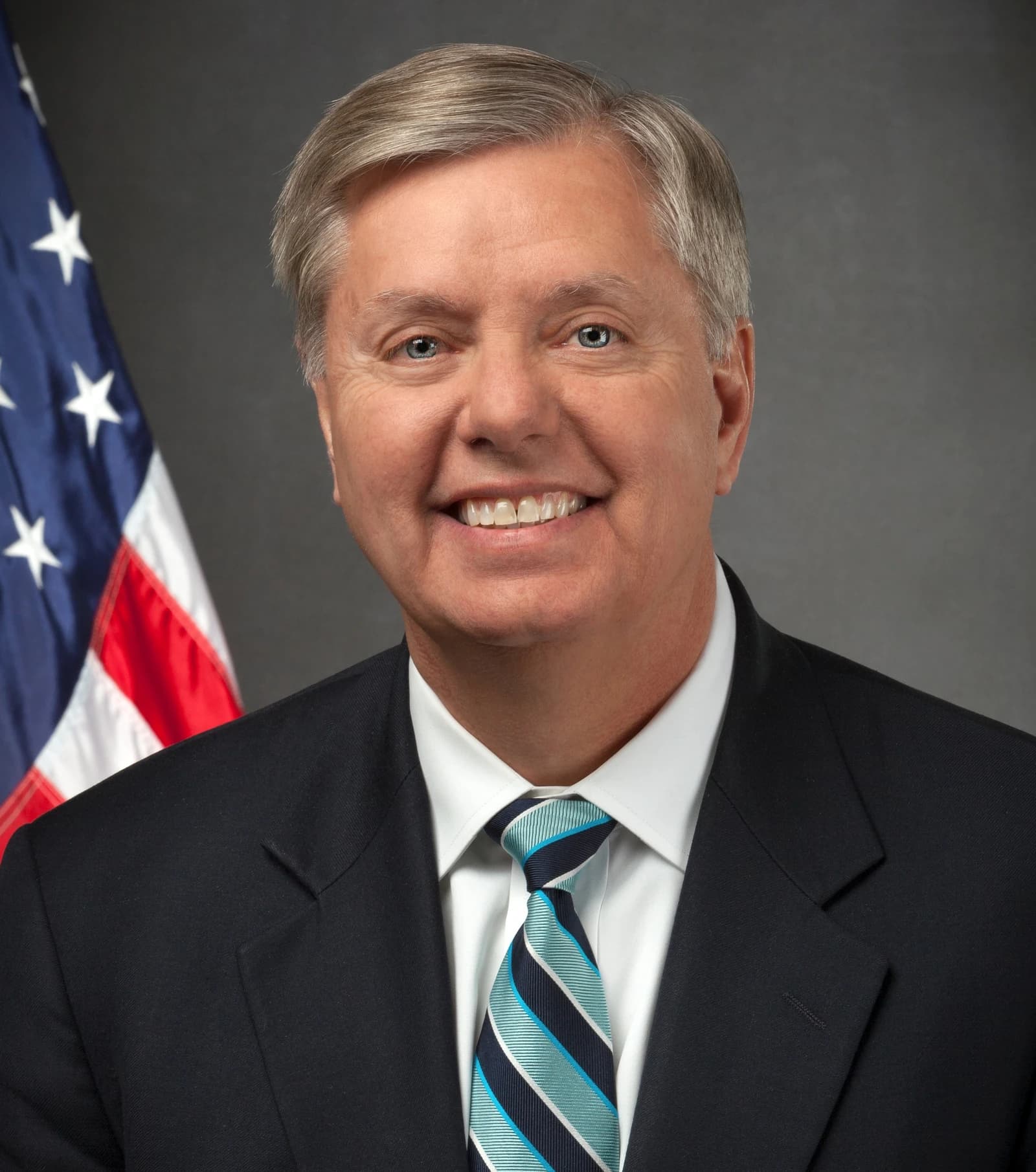 Lindsey Graham, U.S. Senator, smiles in an official portrait with an American flag behind him.