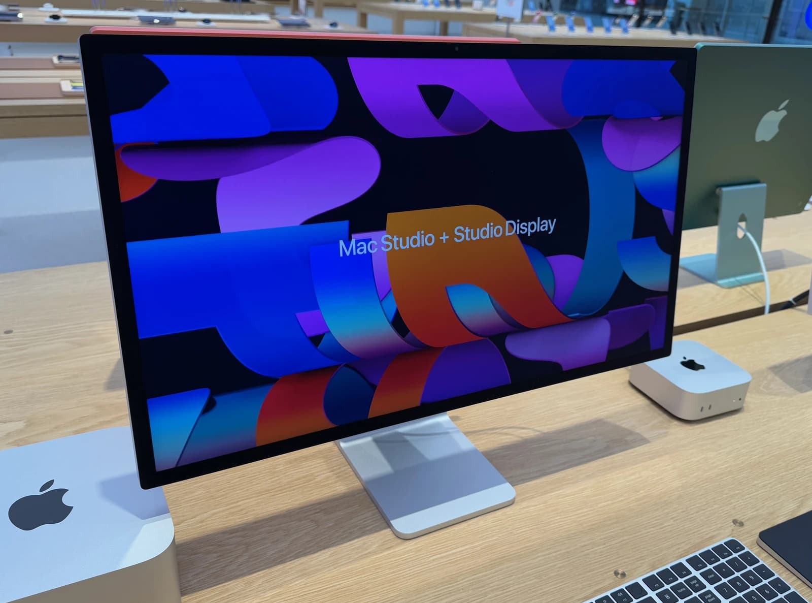 Apple Studio Display showing promotional graphics, with Mac Studio computers nearby.