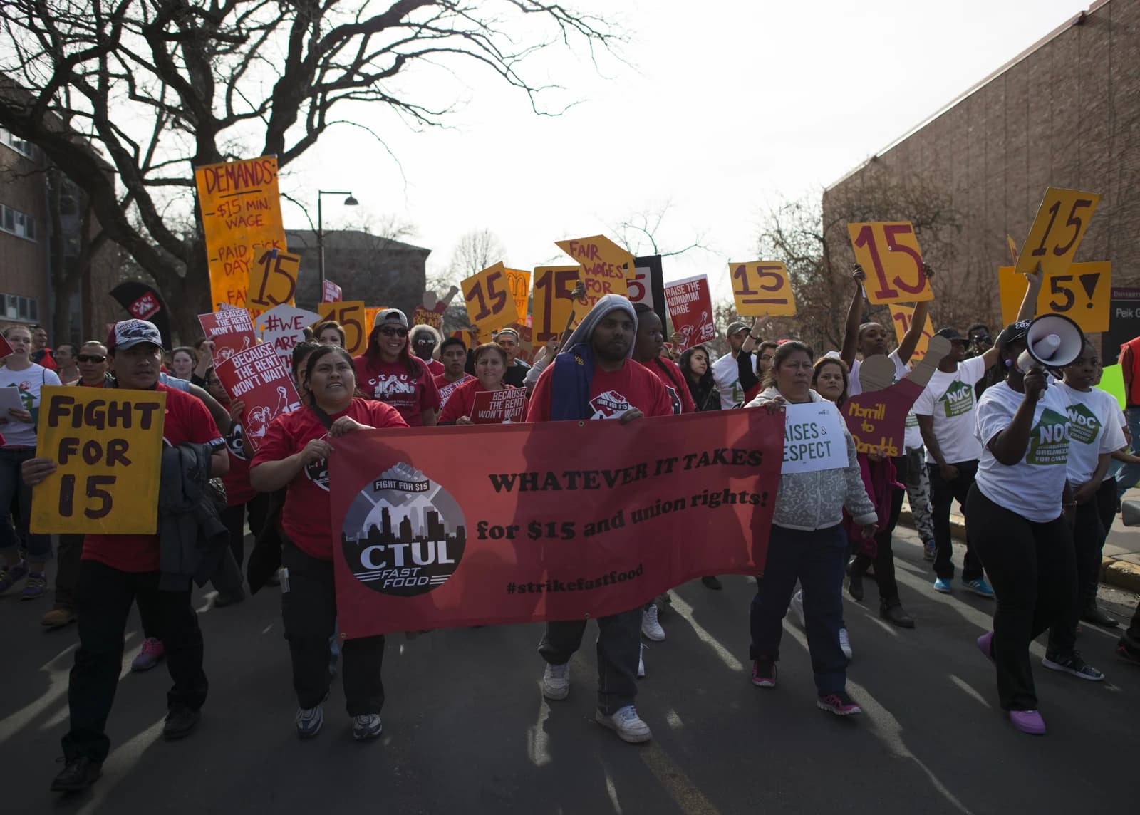 People march down a street holding signs and a banner demanding a minimum wage increase.