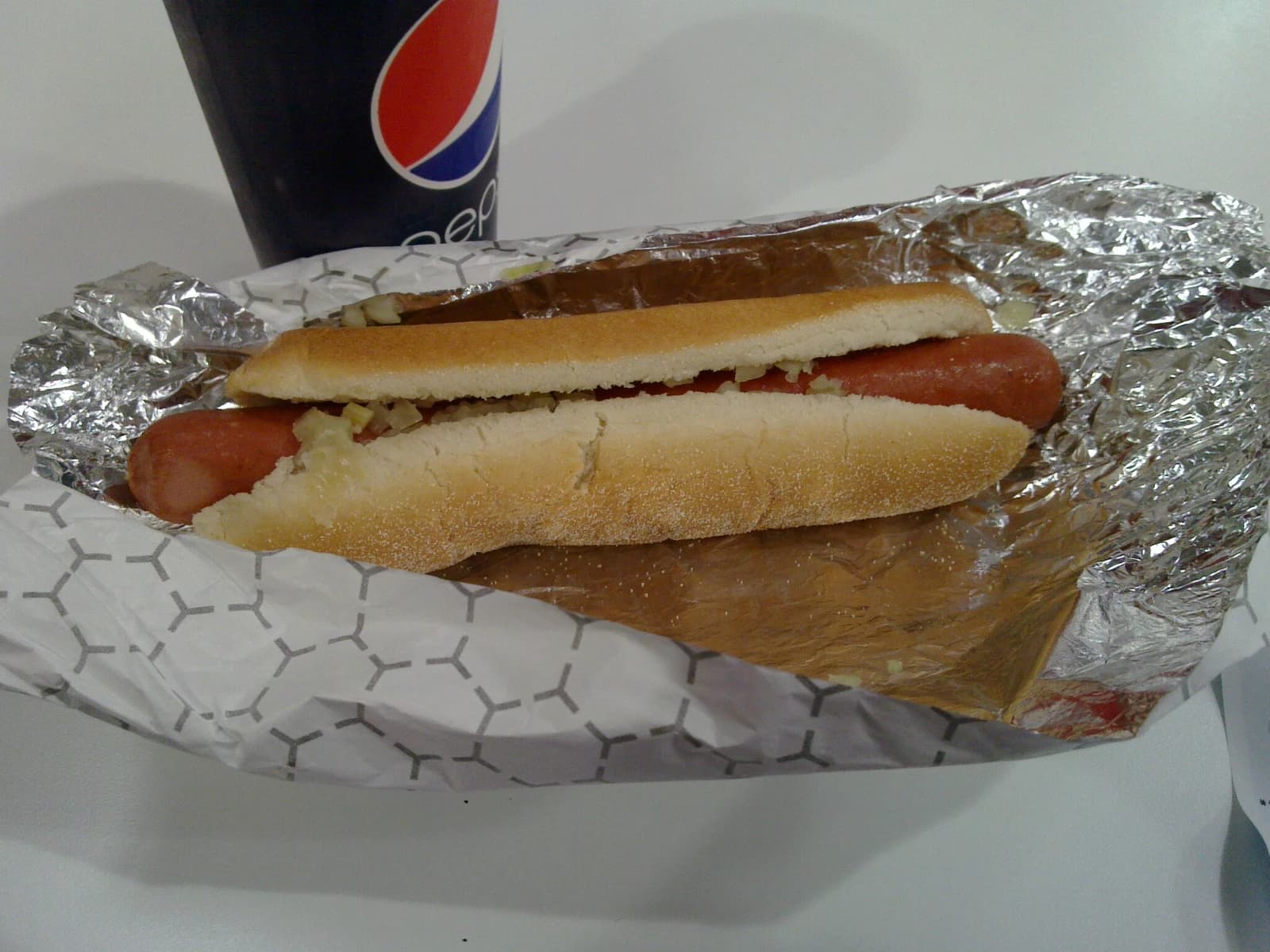 A Costco hot dog in a bun with toppings, wrapped in foil, next to a soda cup.