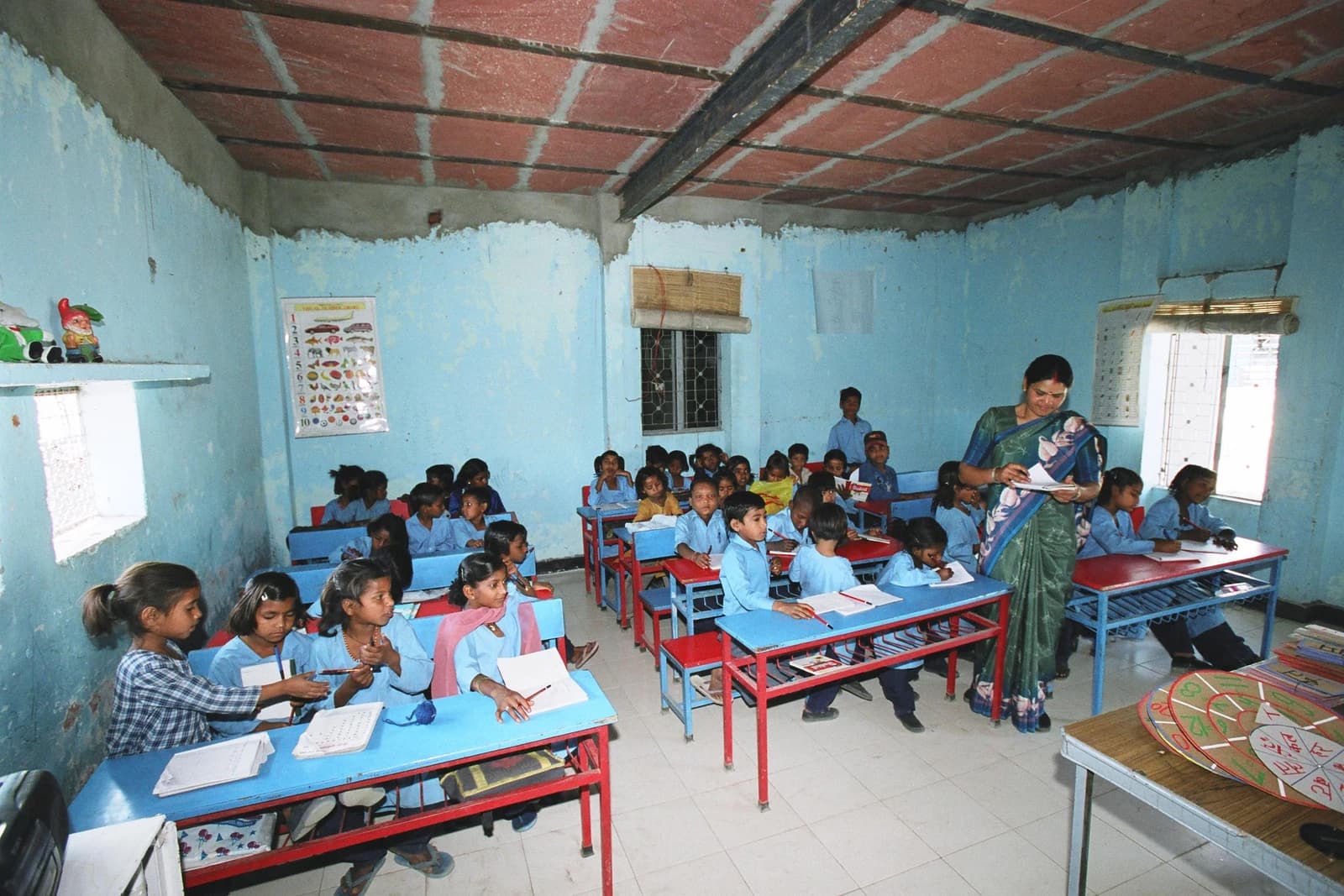 Children in school uniforms sit at desks in a classroom, with a teacher standing.