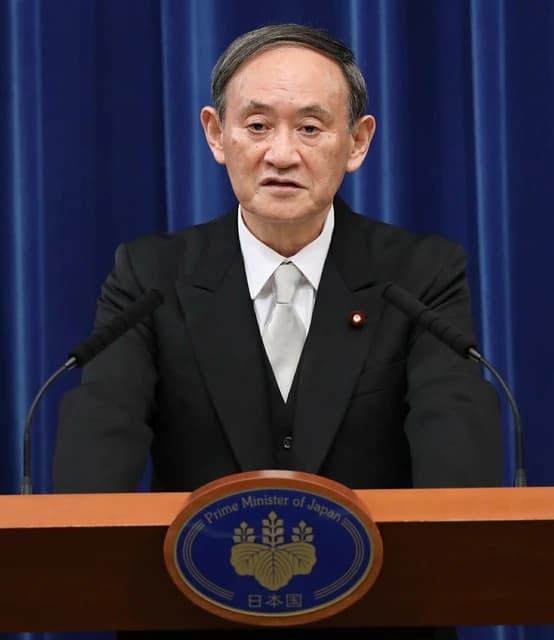 Yoshihide Suga, former Prime Minister of Japan, standing at a podium with microphones.