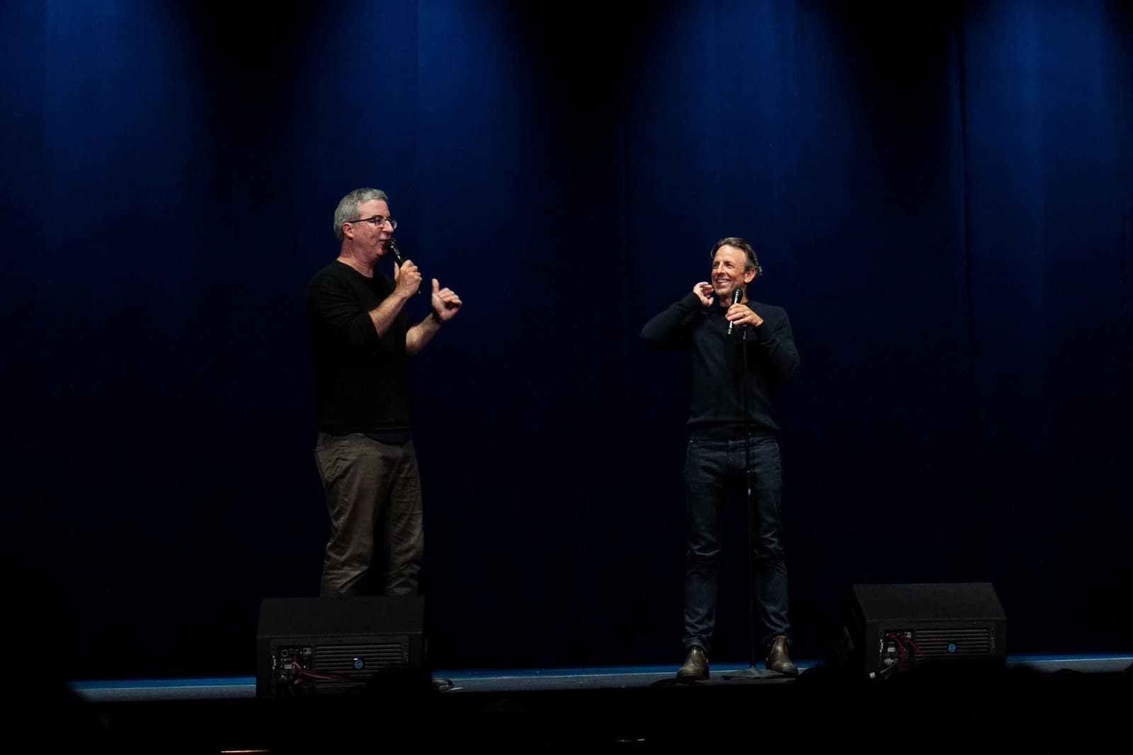John Oliver and Seth Meyers speaking on stage with microphones in hand.