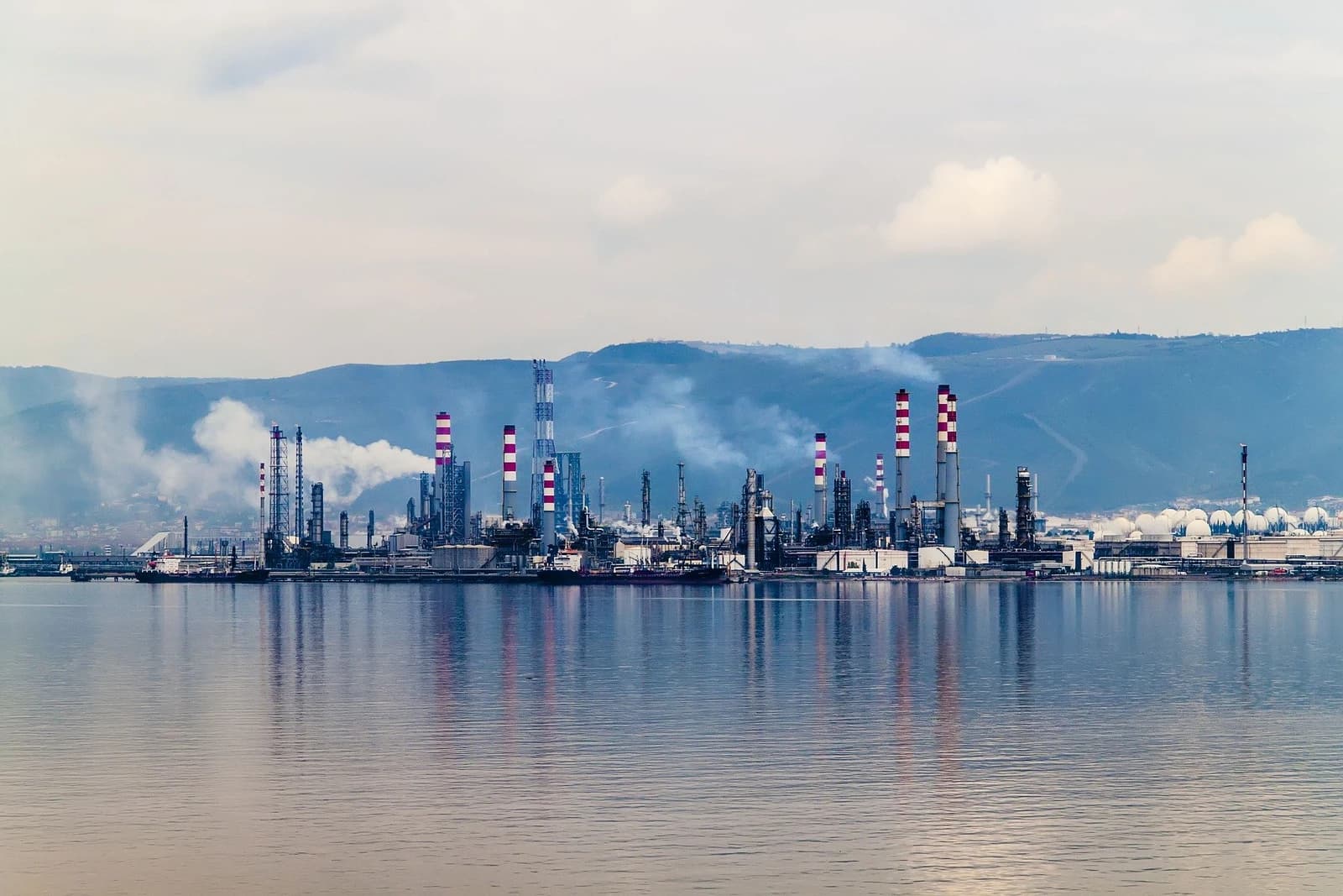 Industrial oil refinery complex with many smokestacks and storage tanks along a body of water.