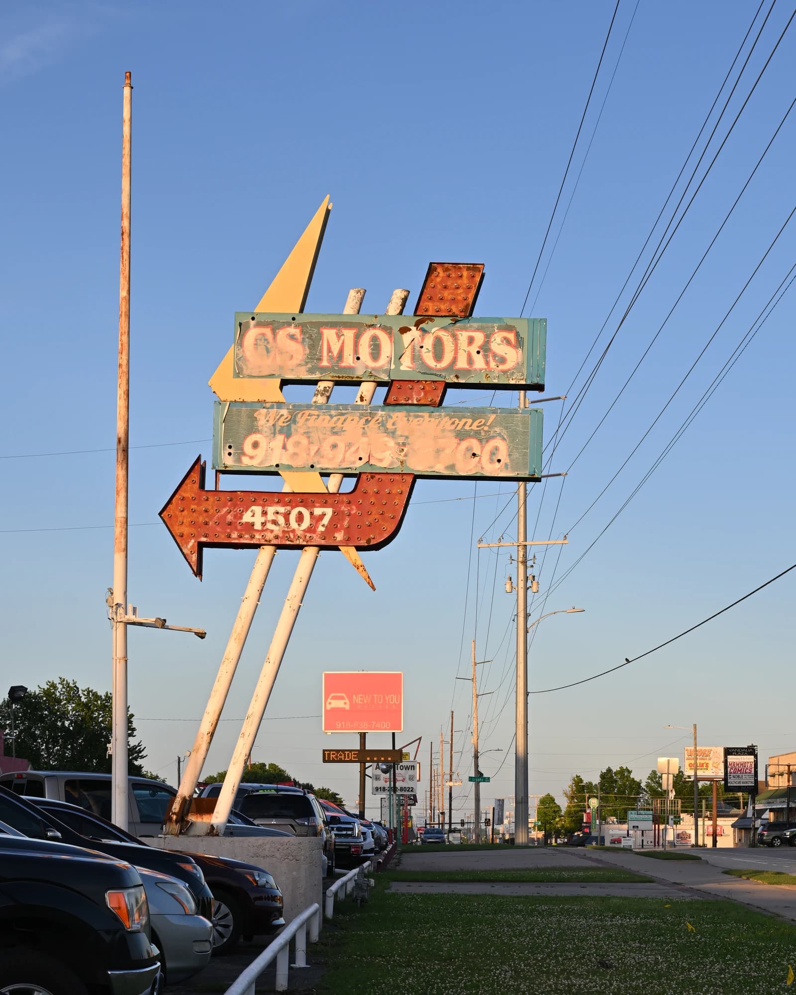 An old, weathered sign for CS Motors car dealership in Tulsa, Oklahoma, with cars parked along the street.