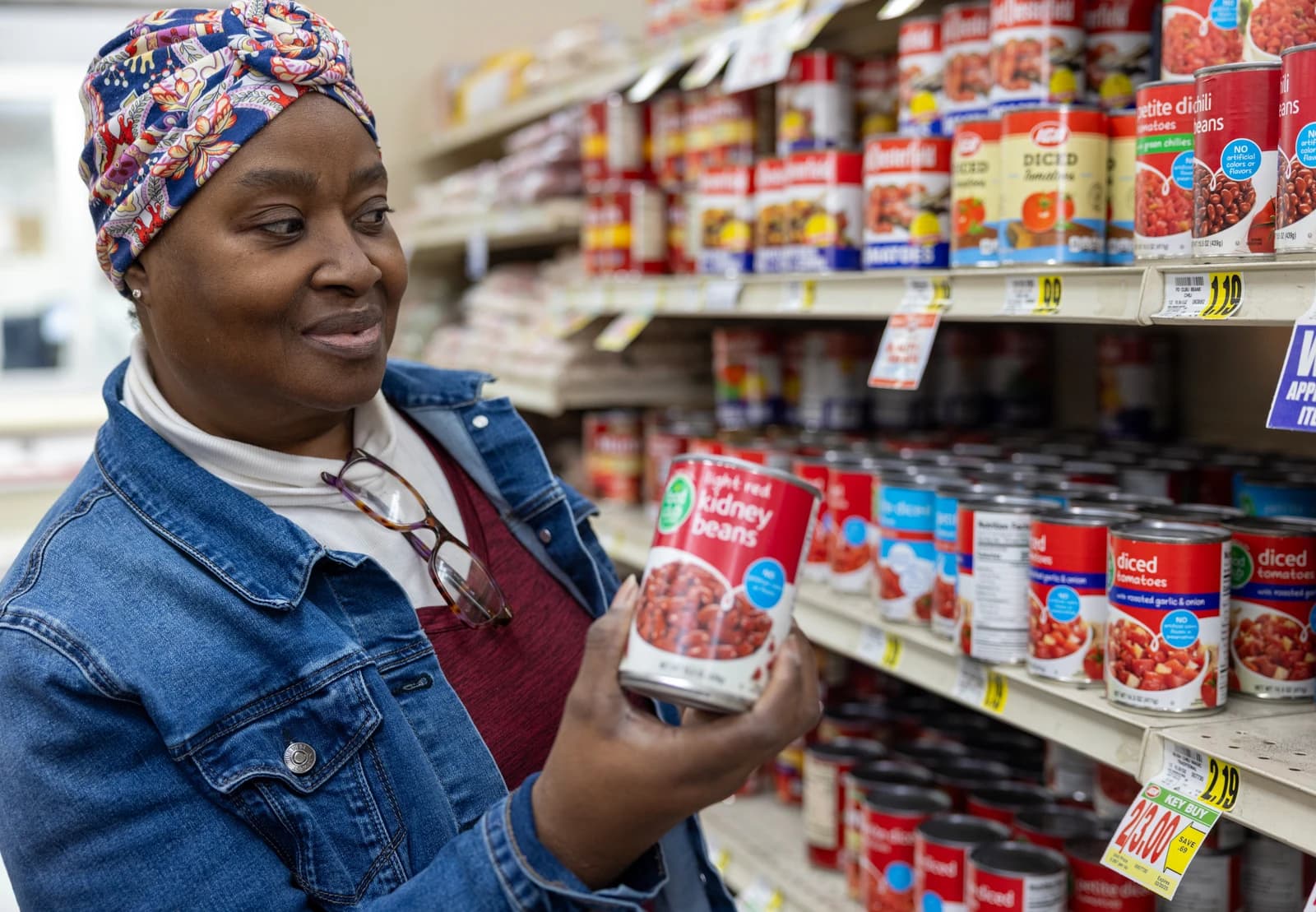 Woman wearing a colorful head covering holds a can of red kidney beans in a grocery store aisle.