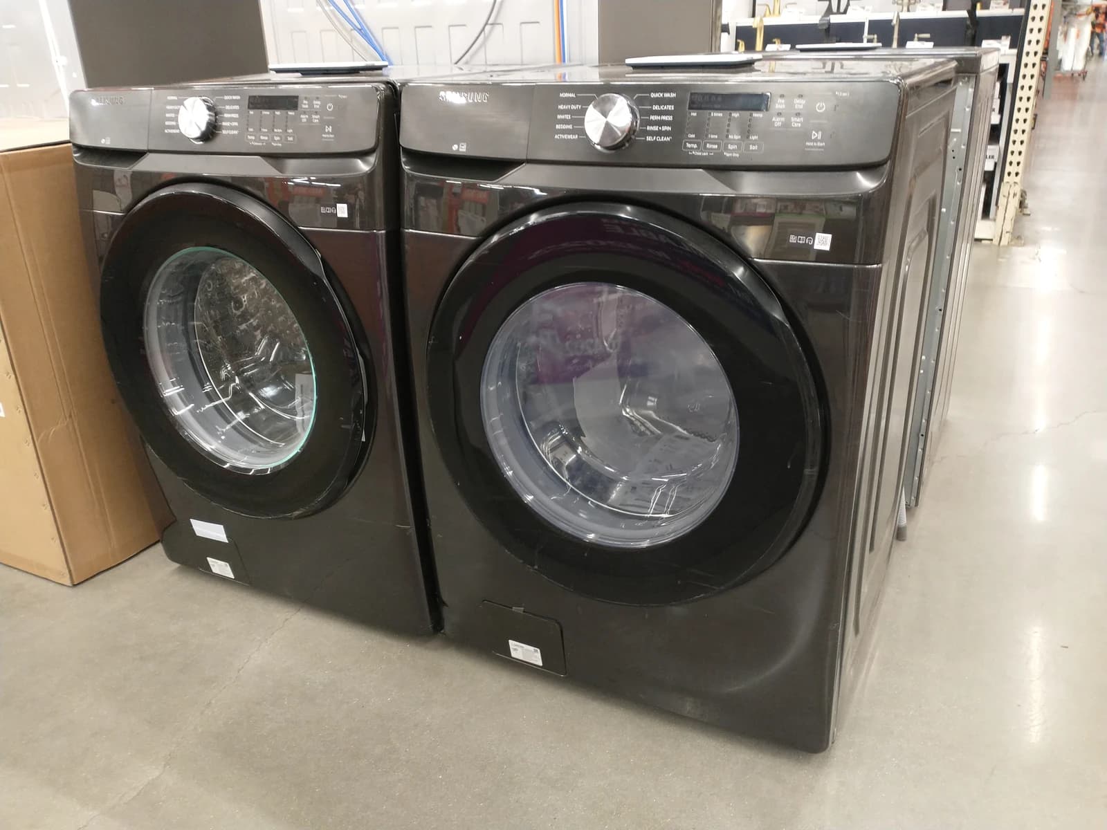 Two black front-load washing machines with controls on top, displayed in a store aisle.