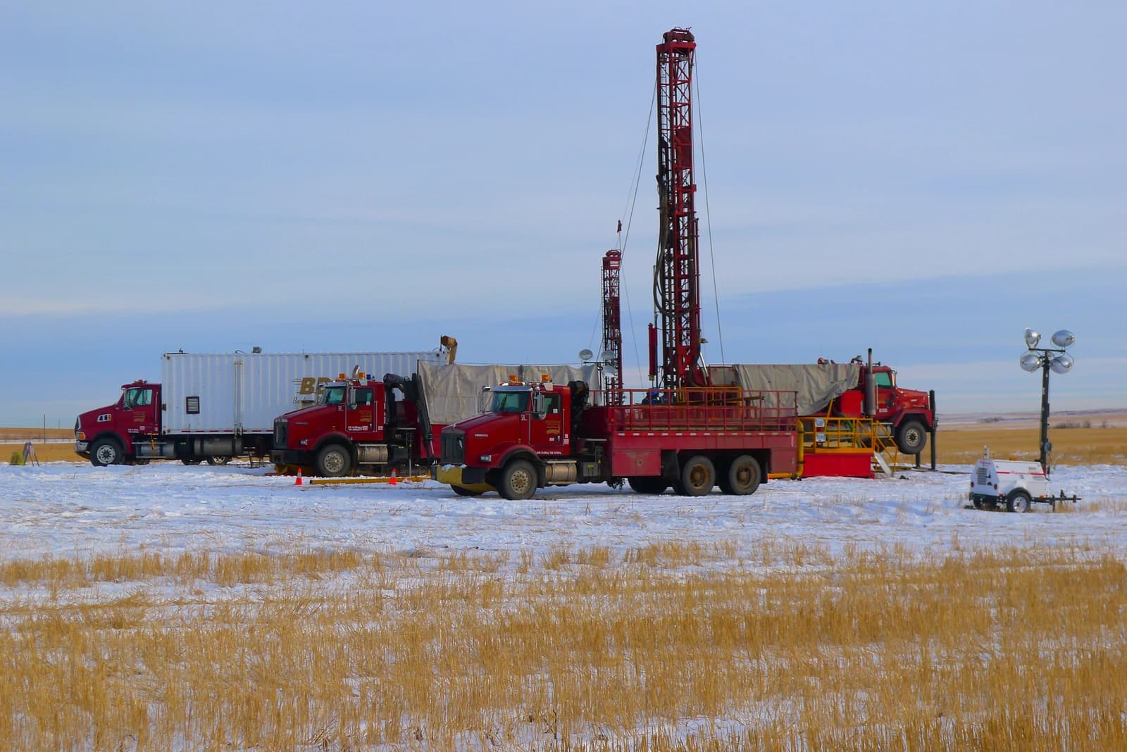 Oil drilling rig and red industrial trucks in a snow-covered field under a cloudy sky.