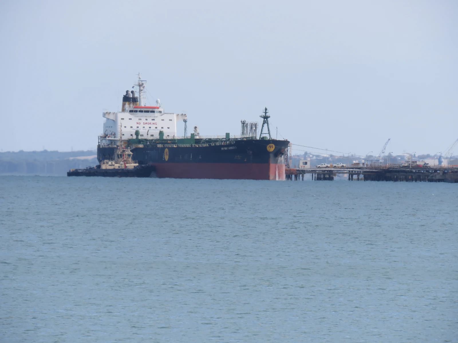 An oil products tanker is docked at a jetty next to a refinery, with a tugboat alongside.