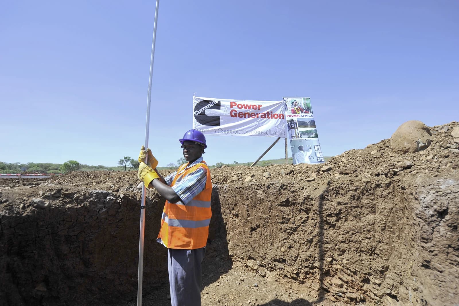 Construction worker wearing hard hat and safety vest, holding measuring pole at a power generation site.