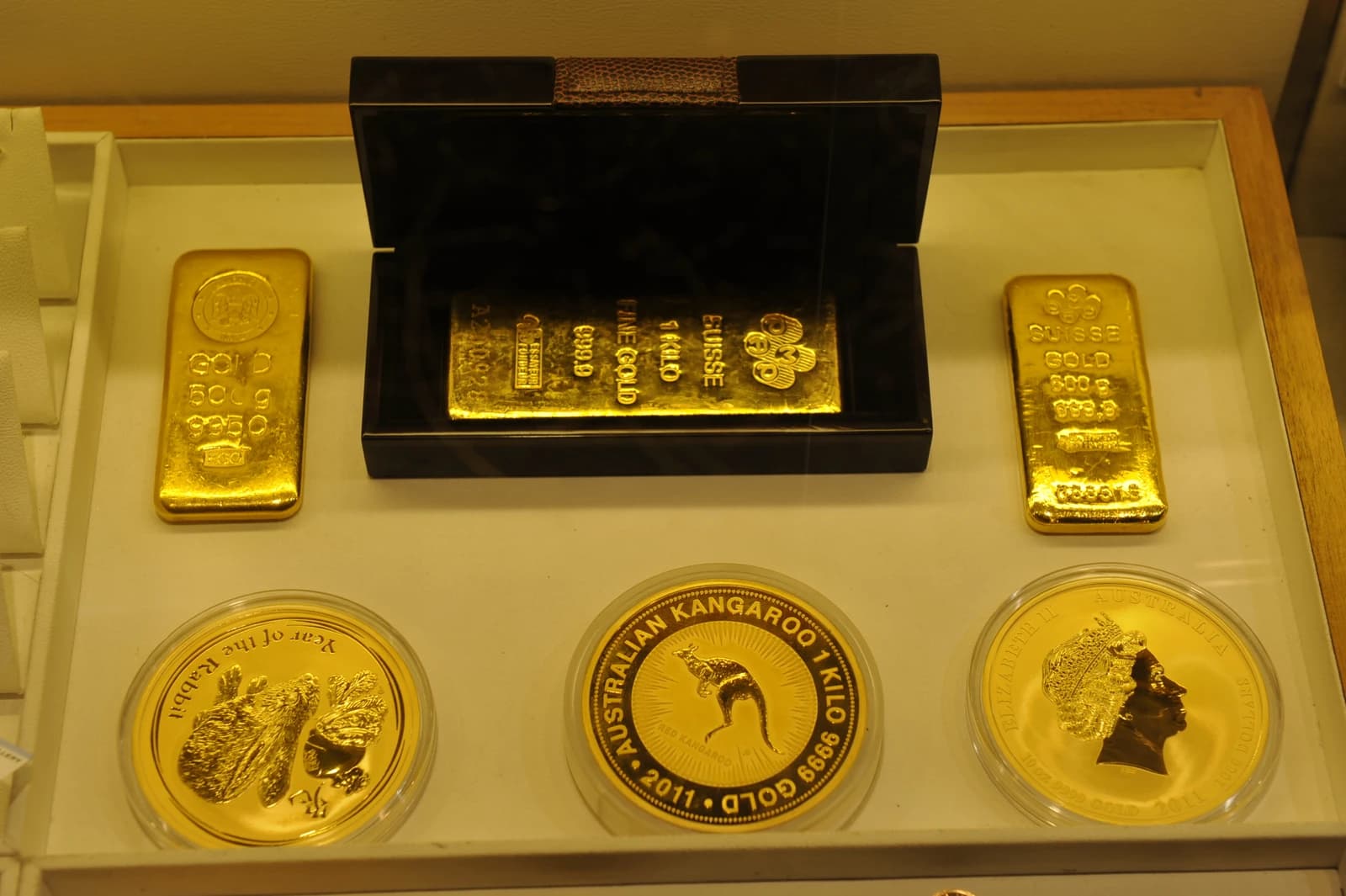 Gold bars and three different gold coins displayed in a glass case.