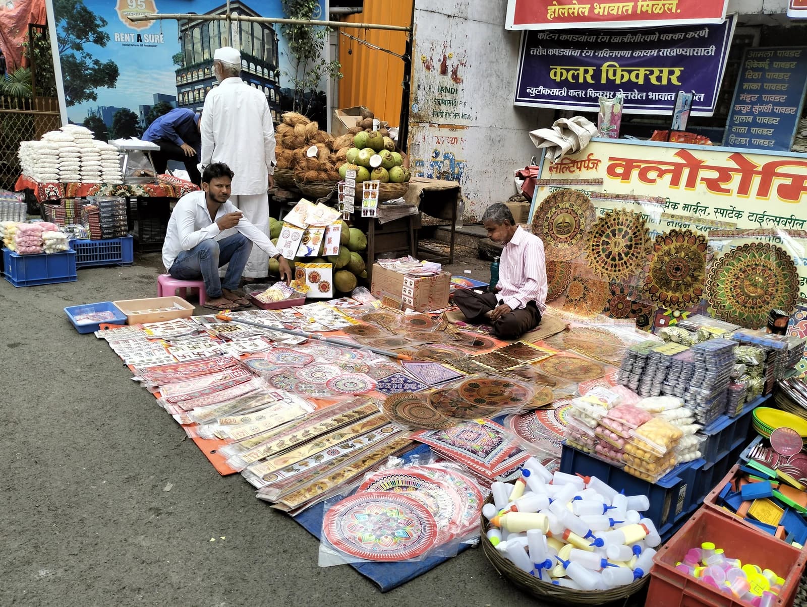 Two men sit on the ground next to a display of various items for sale on a street.
