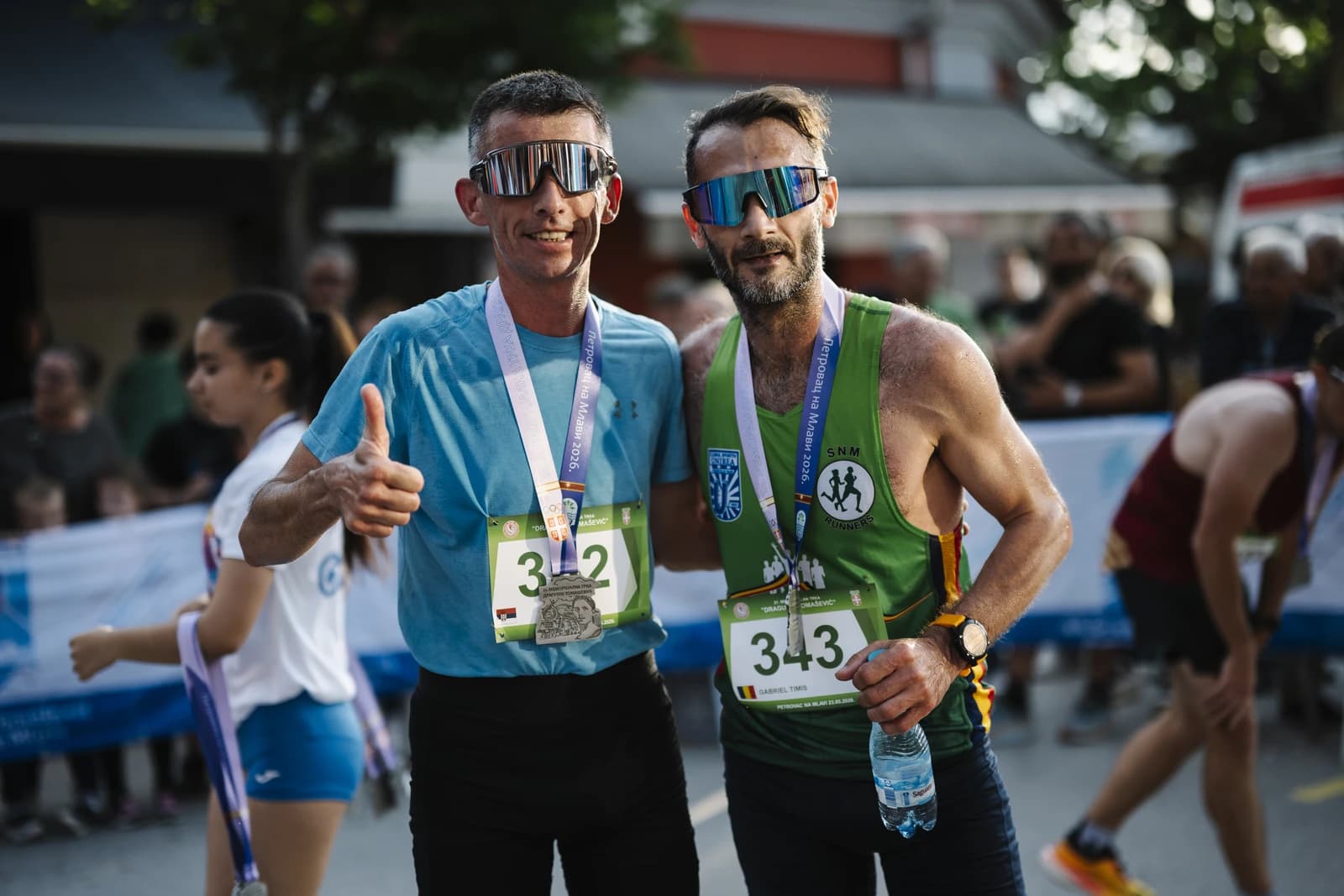 Two male runners wearing race medals and sunglasses pose for a photo.