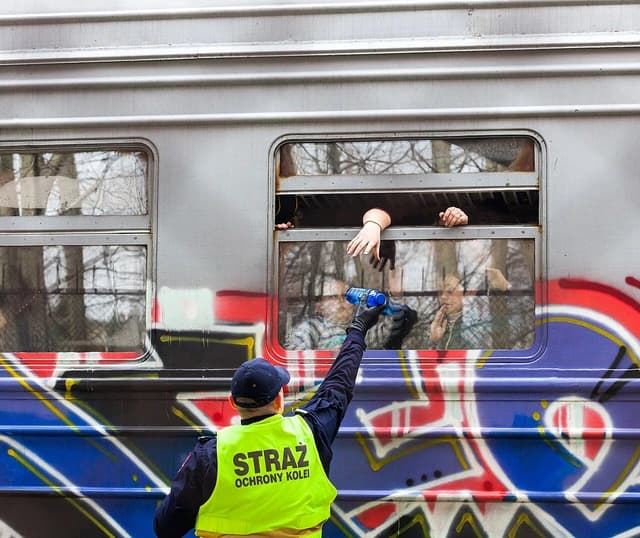 Polish railway guard hands a bottle of water to outstretched hands from a train window.