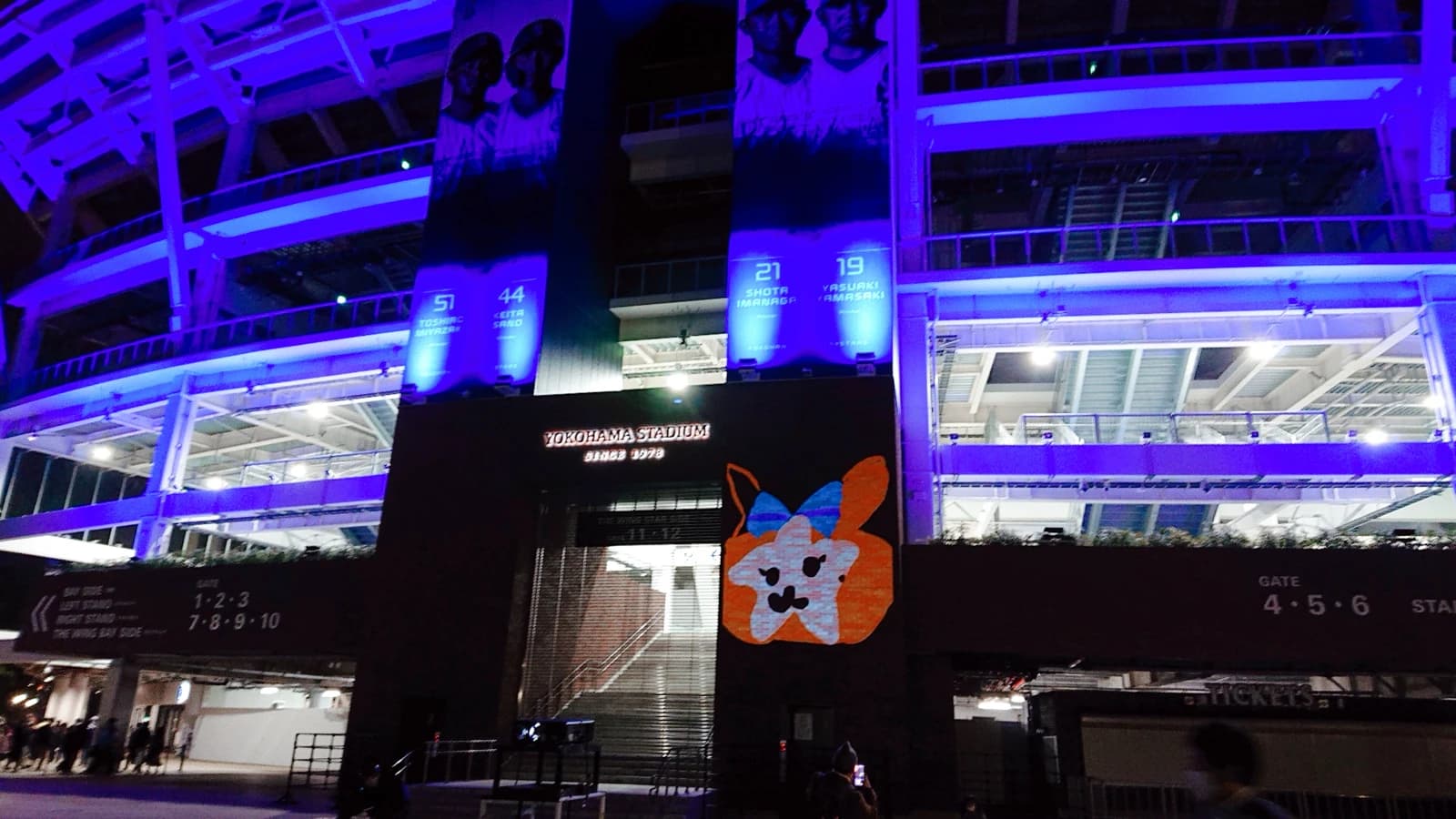 Exterior of Yokohama Stadium at night with prominent player banners and purple lighting.
