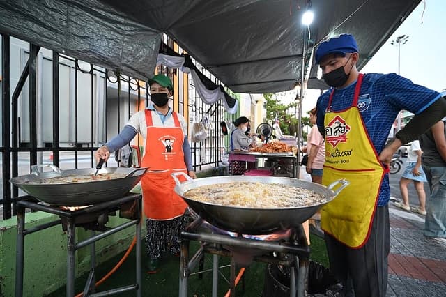 Two street vendors cook food in large woks at an outdoor market stall.