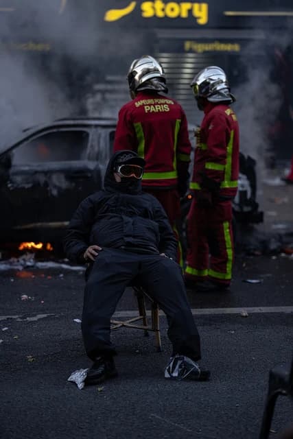 Masked man sits on a chair, firefighters stand behind a burning car.