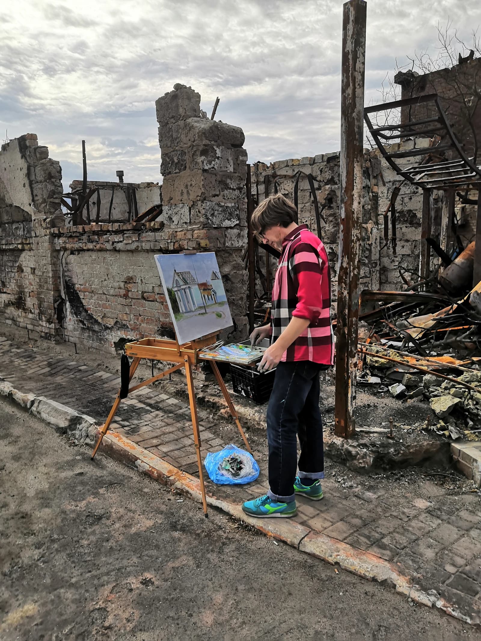 An artist painting a house stands in front of a ruined building in Irpin, Ukraine.
