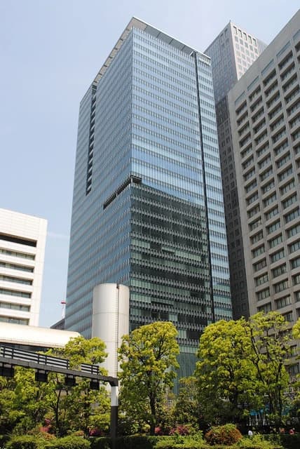 Nikkei Inc. headquarters building, a modern skyscraper with glass facade, surrounded by other buildings and green trees.
