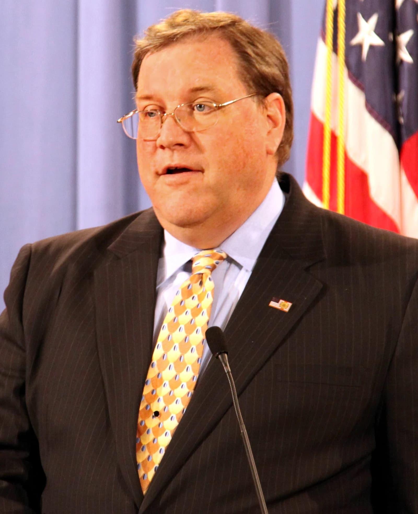 A man wearing glasses and a suit speaks at a microphone during a press conference.