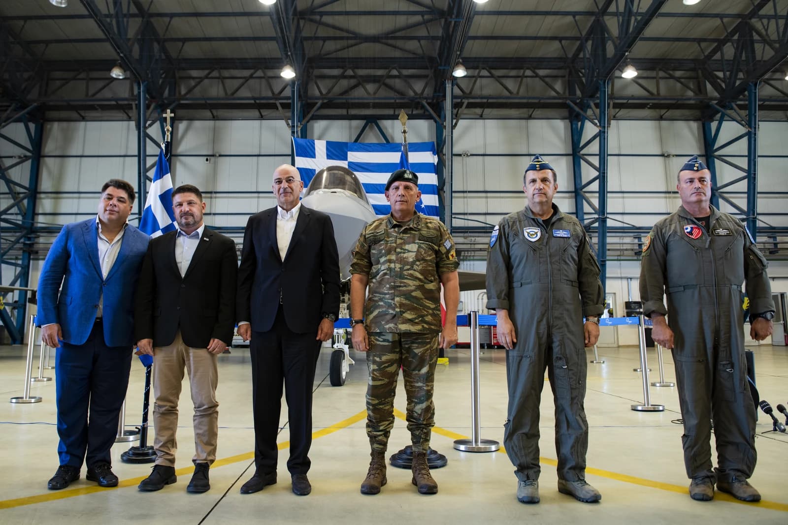 Group of politicians and military leaders posing in front of a Greek flag and aircraft.