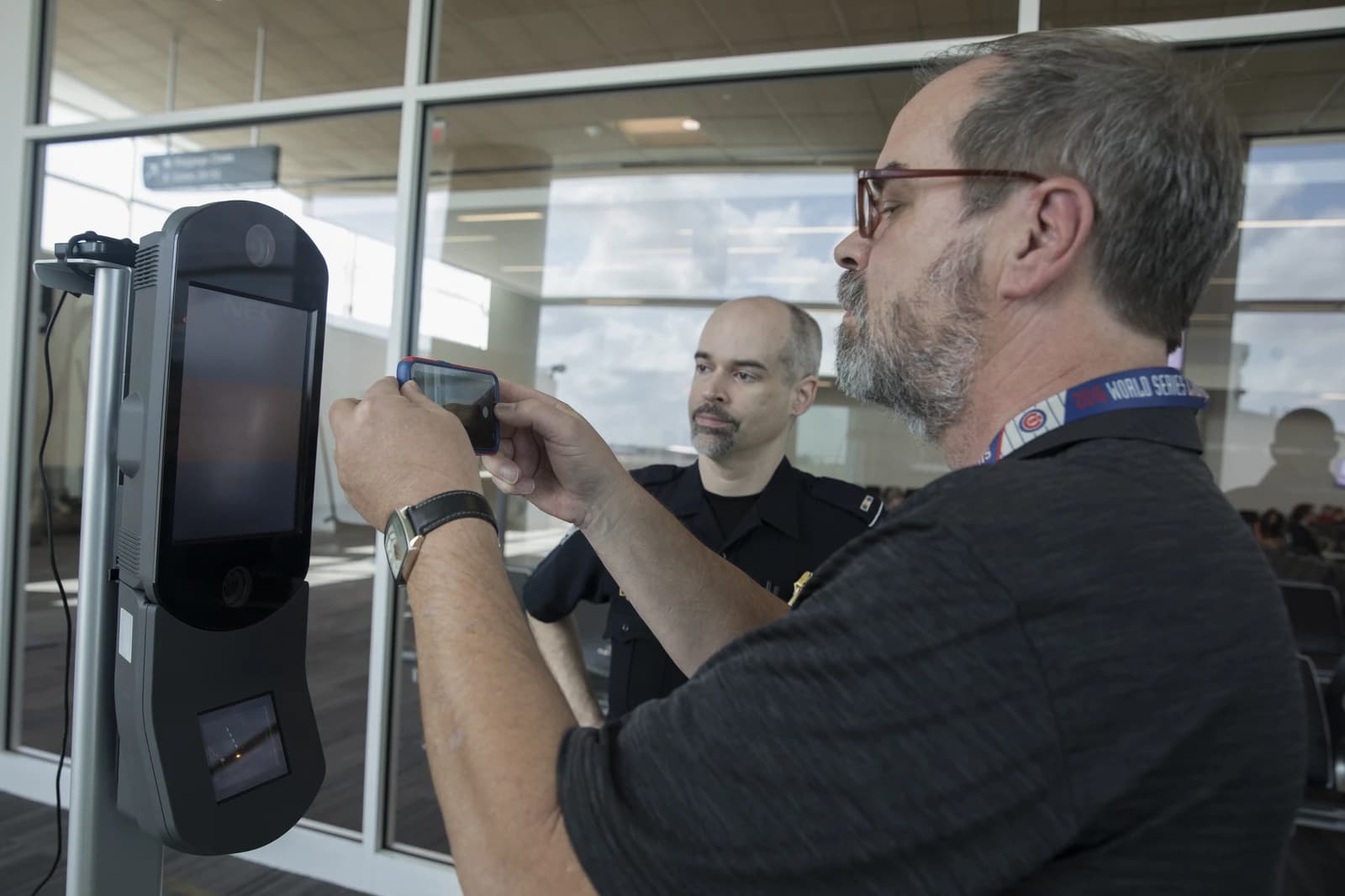 Man holding a smartphone in front of a biometric processing terminal at an airport.