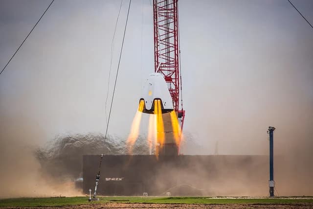 SpaceX Dragon 2 spacecraft suspended by a crane during a hover test, engines firing.