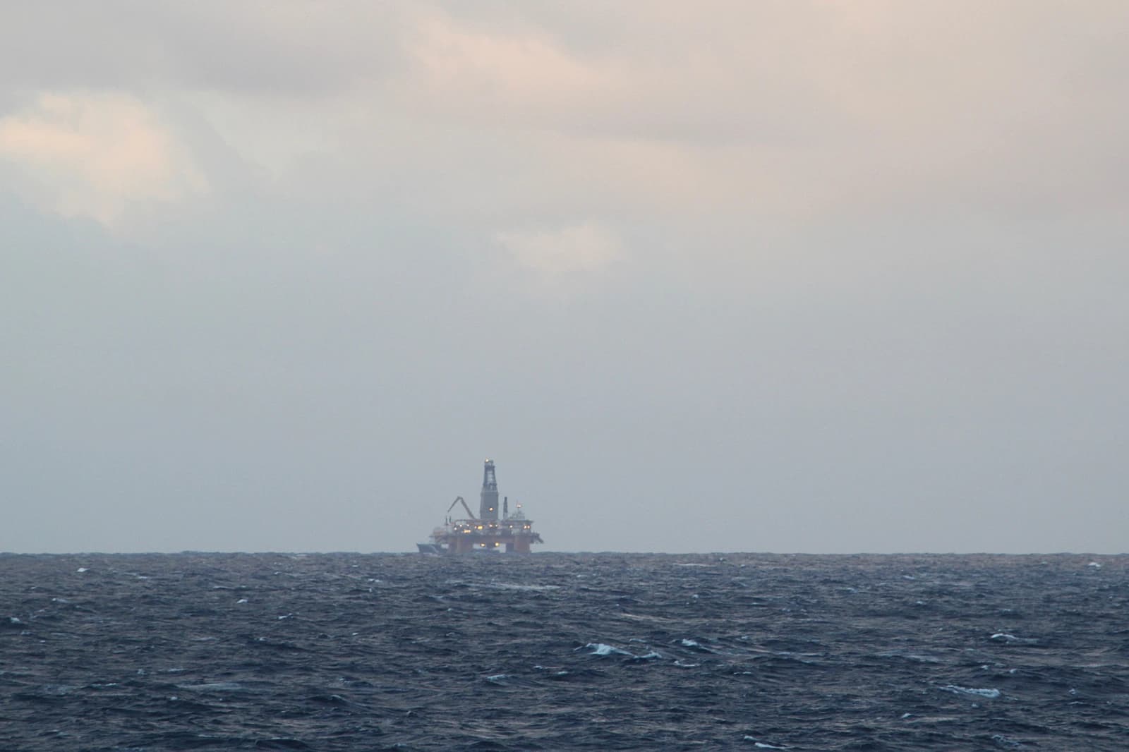 An offshore oil platform visible in the distance across choppy dark blue ocean water under a grey sky.
