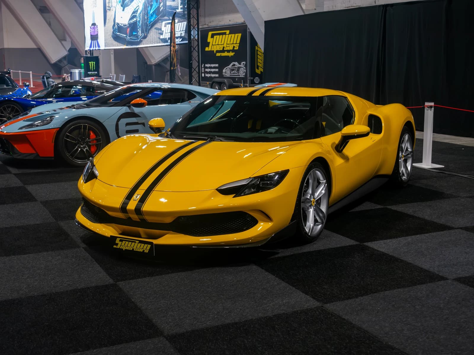 Yellow Ferrari 296 GTB sports car on display indoors with black racing stripes.