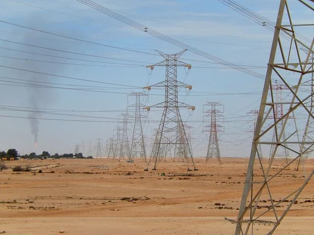 Numerous electrical power lines stretching across a desert landscape in Qatar, with a distant gas flare.