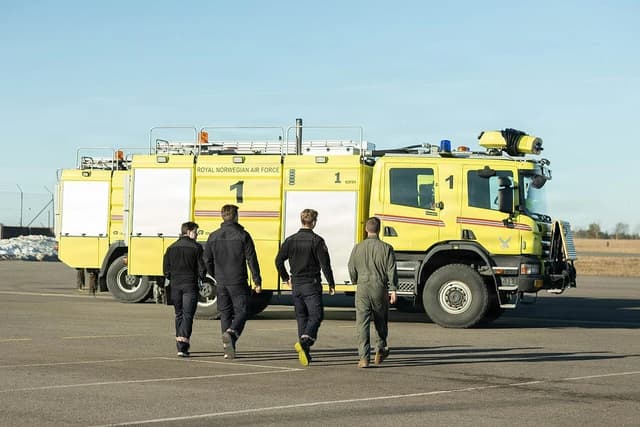 Four airmen walk on an airfield towards a large yellow military vehicle.