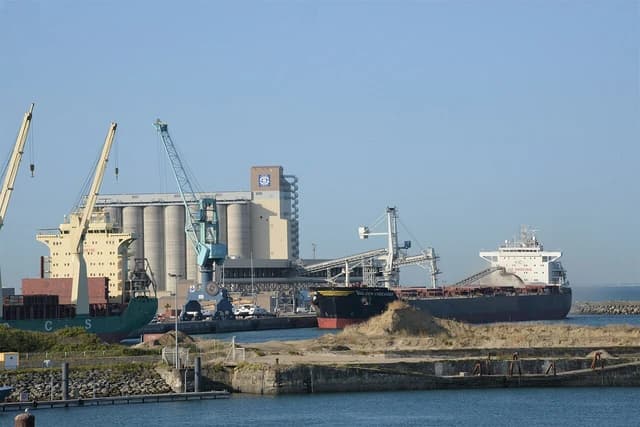 Several large bulk carrier ships docked at an industrial port with grain silos and cranes.