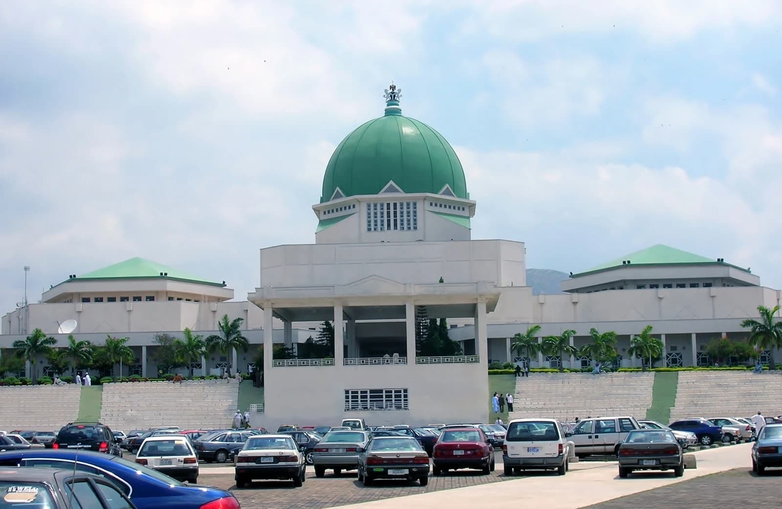 The large green-domed National Assembly building in Nigeria, with a parking lot in the foreground.
