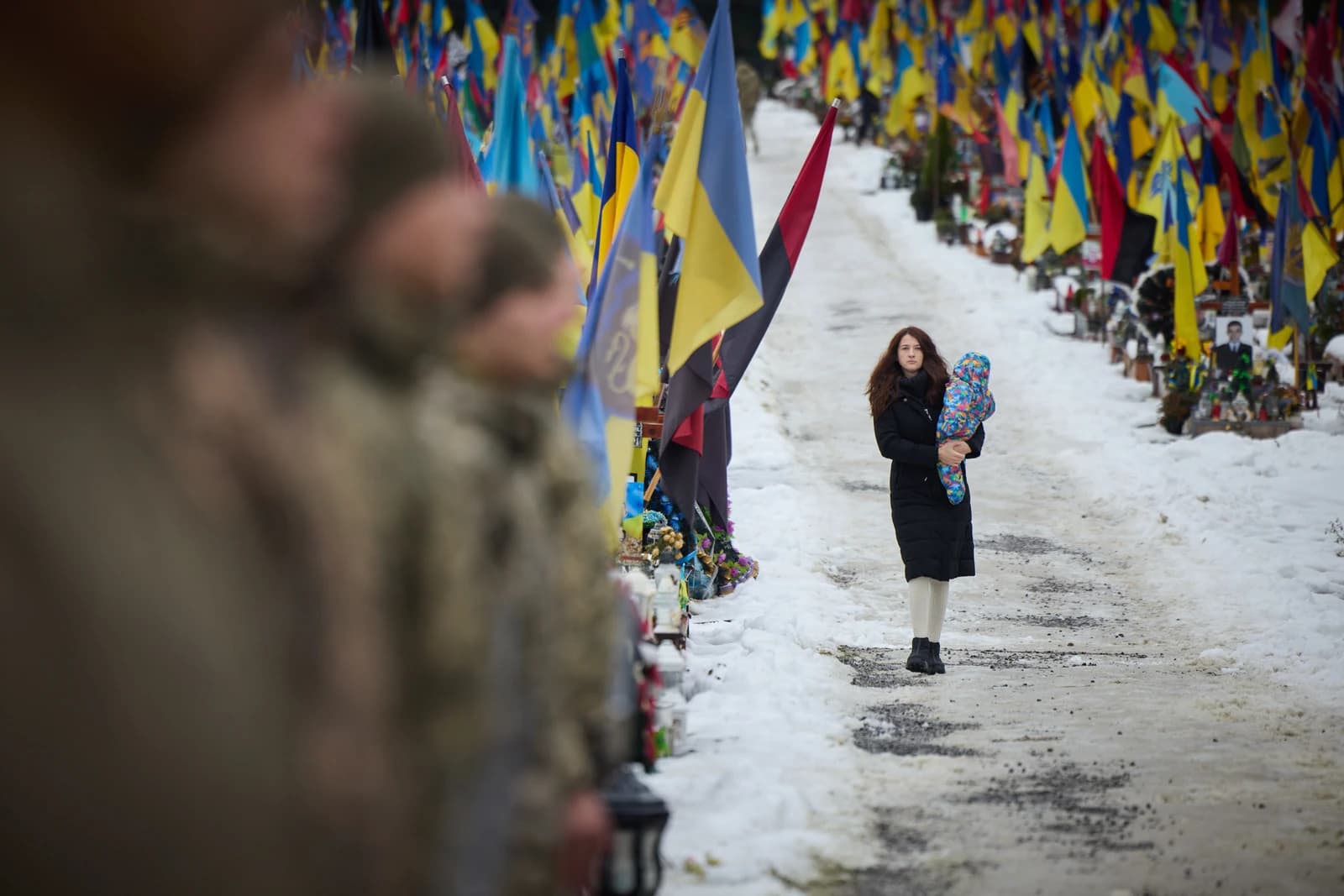 Woman holding child walks past rows of graves with Ukrainian flags in a snowy cemetery.