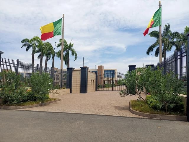 Two flags of Benin flying high on flagpoles at the entrance to a building.