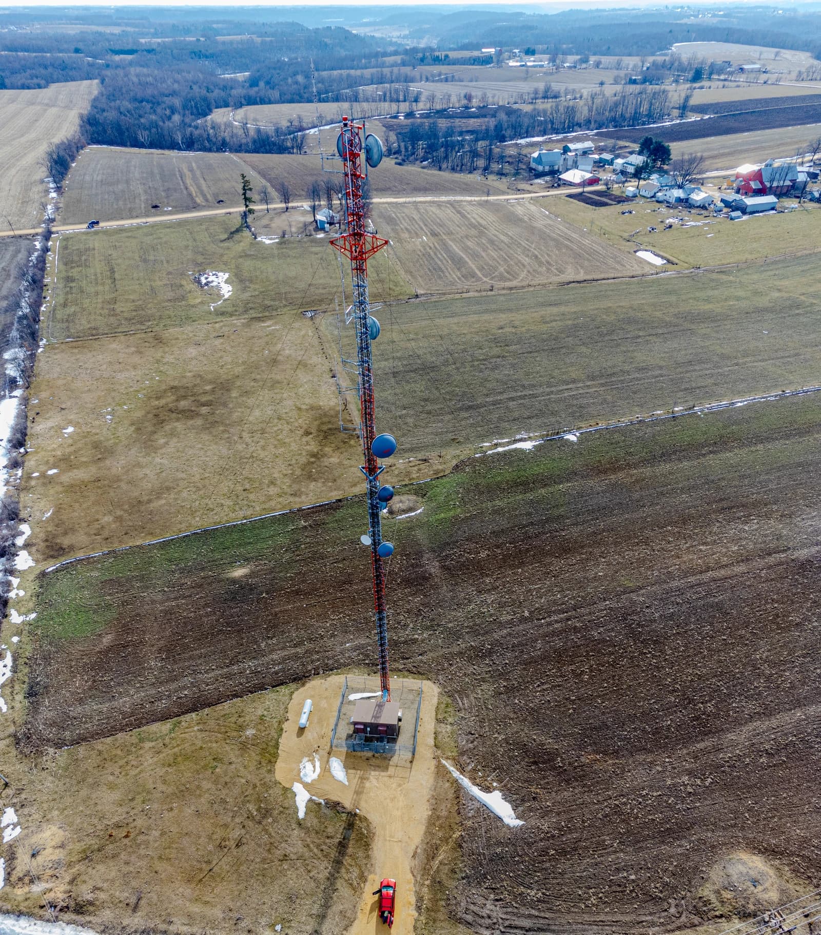 Aerial view of a red and white cell tower with multiple dishes in a field.