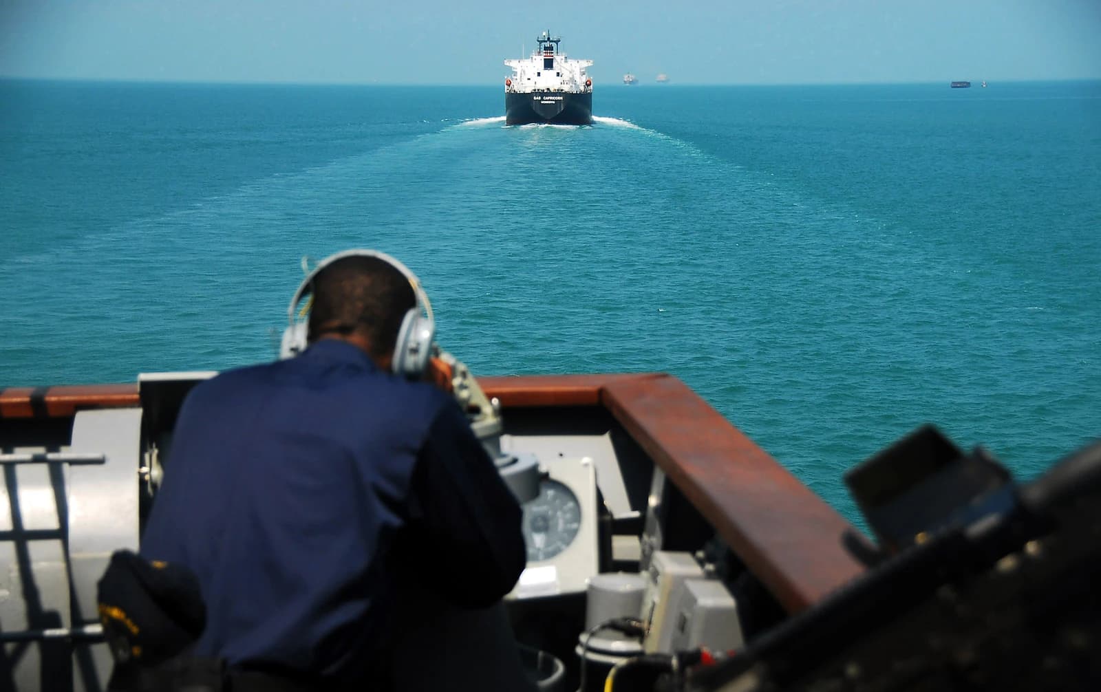 A U.S. Navy Sailor in headphones observes distant ships from a cruiser's deck.