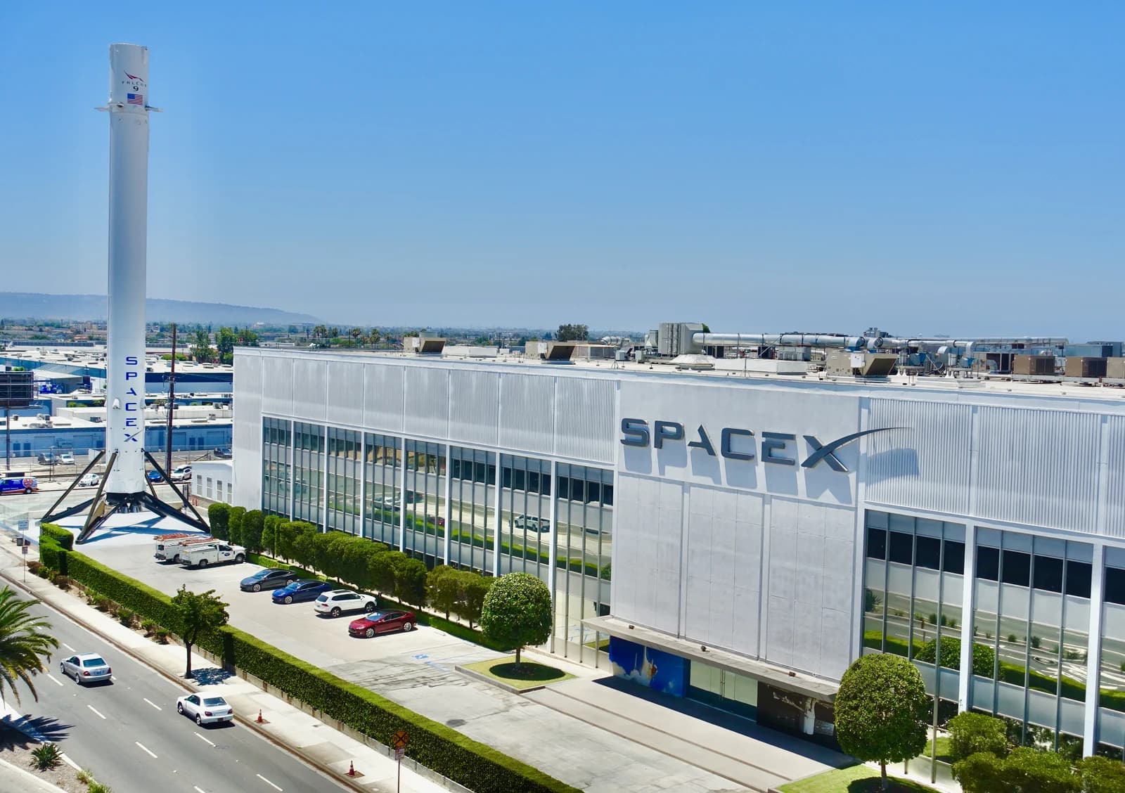 SpaceX factory building with a Falcon 9 rocket booster displayed outside on a sunny day.