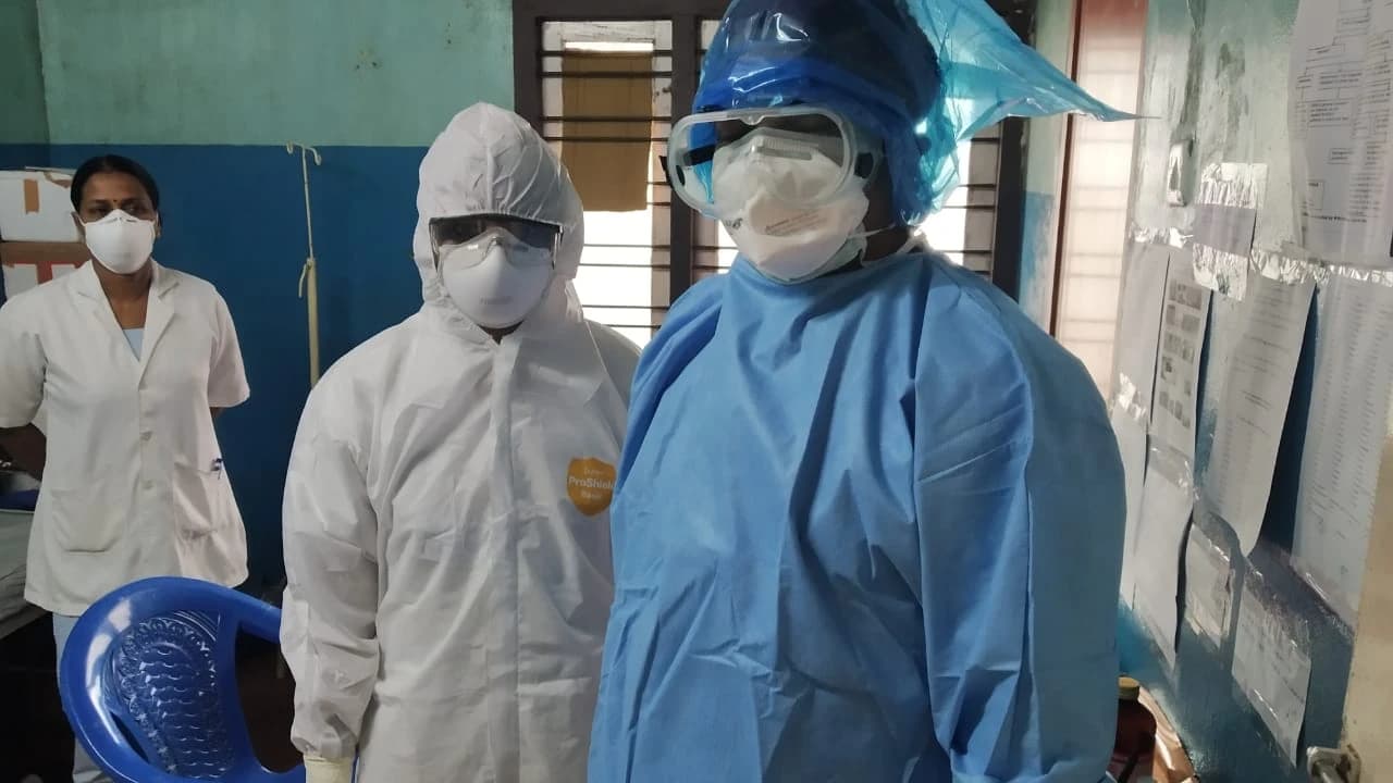 Three healthcare workers, two wearing full PPE and one in scrubs and a mask, standing in a room.