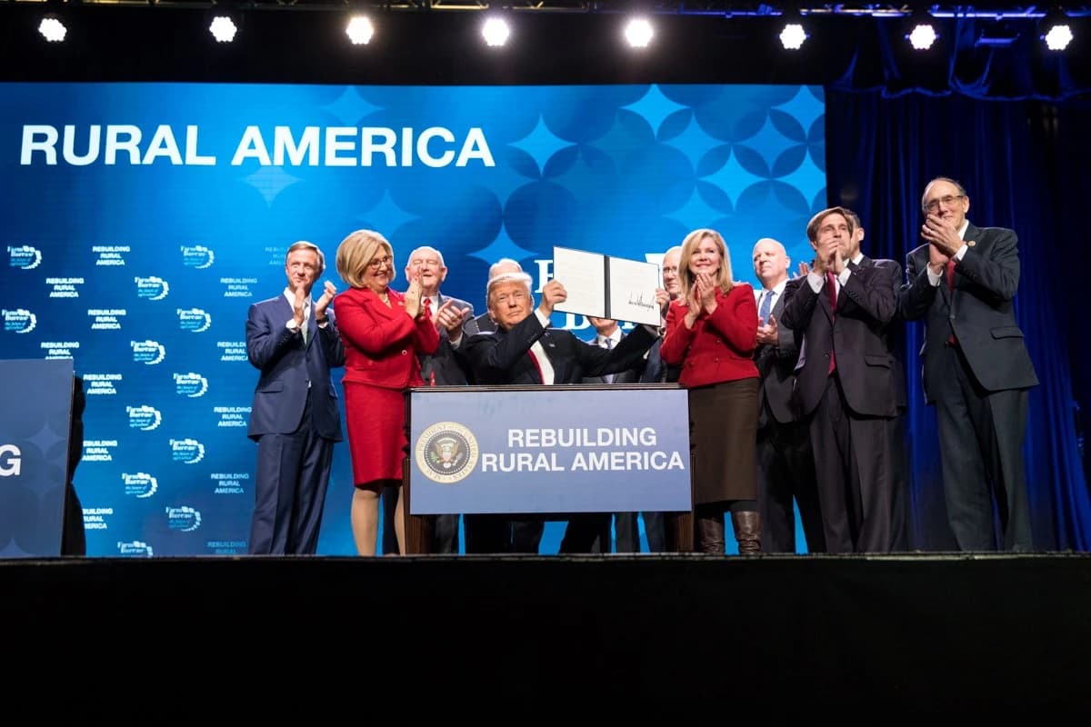 Donald Trump, President, holds up a signed document on stage surrounded by officials.