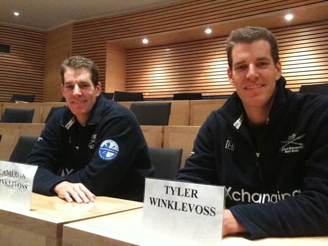 Cameron and Tyler Winklevoss seated at a long table indoors, looking towards the camera.