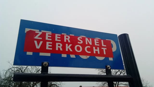 A blue 'sold' sign with red text 'Zeer Snel Verkocht' in a residential area.