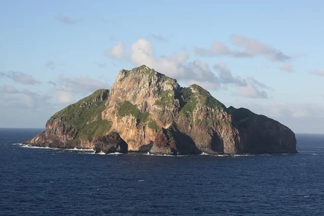 Hunter Island, a rocky, green-capped island, rises from the deep blue ocean under a partly cloudy sky.