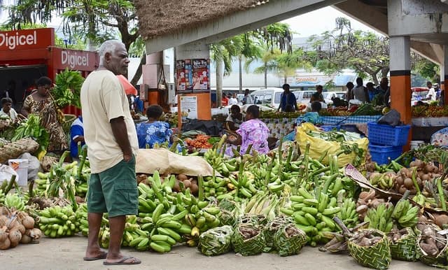 A man stands in a market surrounded by piles of bananas and other agricultural products.